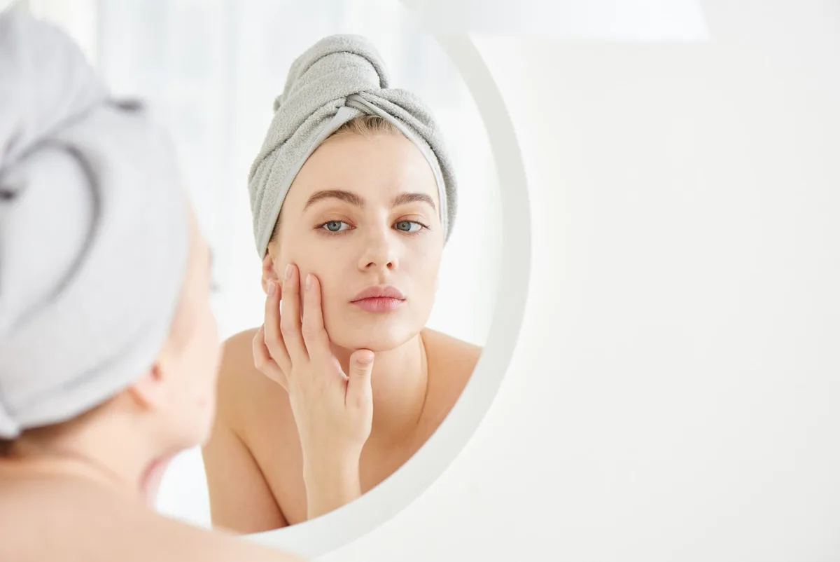 A woman checking her skin in the mirror on day 1 of microneedling aftercare before applying a hyaluronic acid serum.