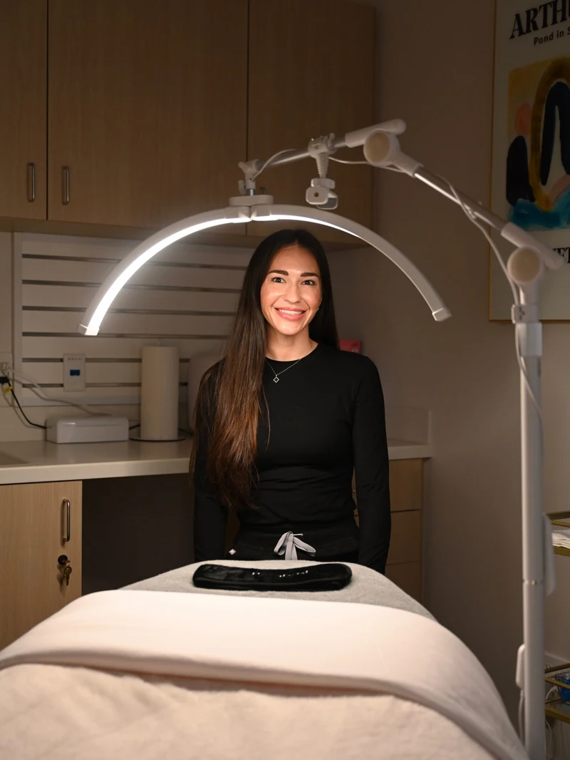 Danielle Hancock, Licensed Aesthetician, seated on the treatment bed under the LED arch lamp in the facial room at Coastal Health & Medical Spa in Jacksonville